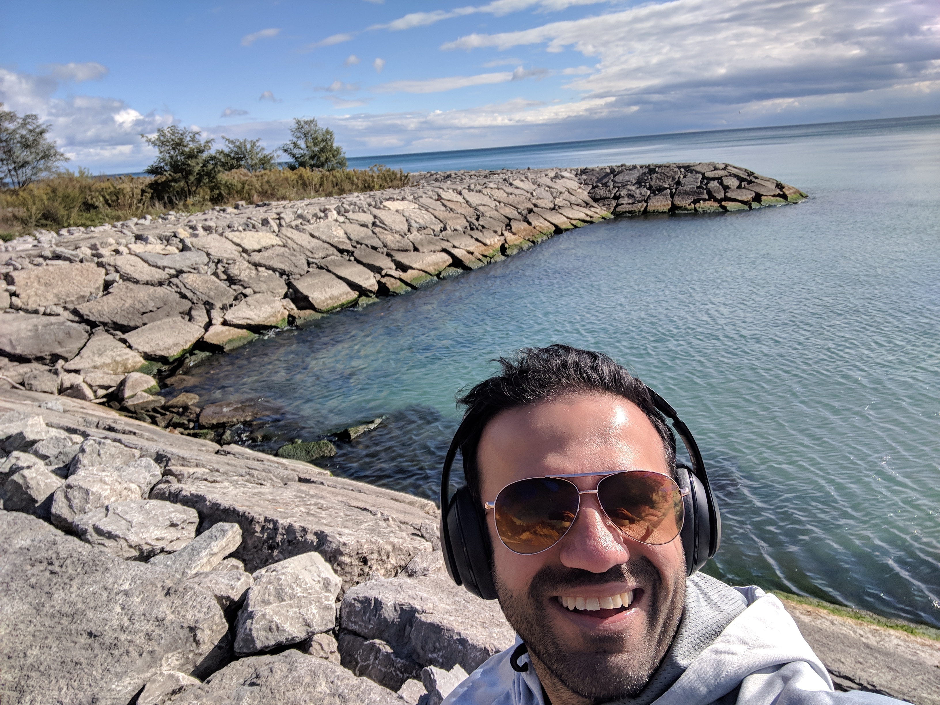 Shabbir Evershine is smiling taking a selfie, wearing sunglasses and has black headphones on his head. He's wearing a grey hoodie sweater. He's standing on rocks at a pier in Toronto. Beautiful blue water surrounds him also with a nice blue and white sky.